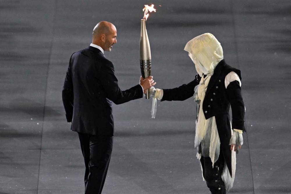 Un portador de la antorcha pasa la llama olímpica al exfutbolista francés Zinedine Zidane durante la ceremonia de apertura de los Juegos Olímpicos de París 2024 en París el 26 de julio de 2024. (Foto de Fabrice COFFRINI / AFP)