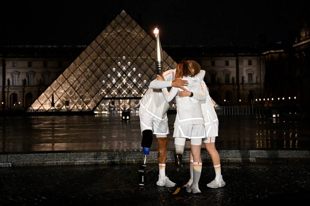 Los atletas franceses y los abanderados paralímpicos de Francia Nantenin Keita y Alexis Hanquinquant y la atleta paralímpica francesa Marie-Amelie Le Fur se abrazan mientras sostienen la llama olímpica durante la ceremonia de apertura de los Juegos Olímpicos de París 2024 en París el 26 de julio de 2024, como el Museo del Louvre. y la Pirámide se ven al fondo. (Foto de Olivier MORIN / AFP)