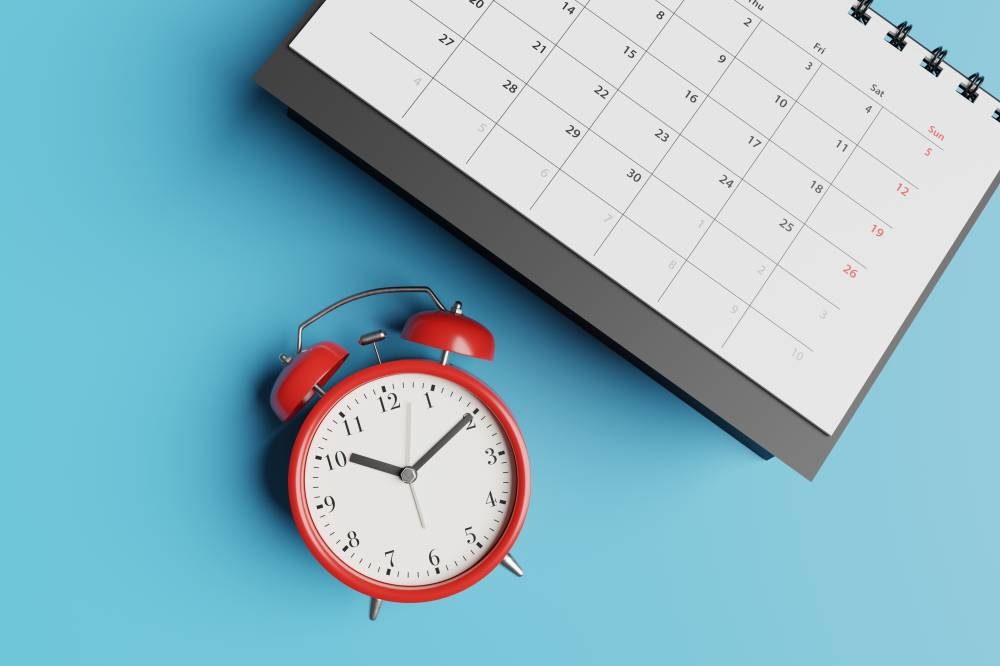 El sector público tiene licencia de vacaciones entre el 1 y el 6 de agosto.,image_description:Black desk calendar and red alarm clock on blue background. Illustration of the concept of tight schedule, deadlines, due dates and workload