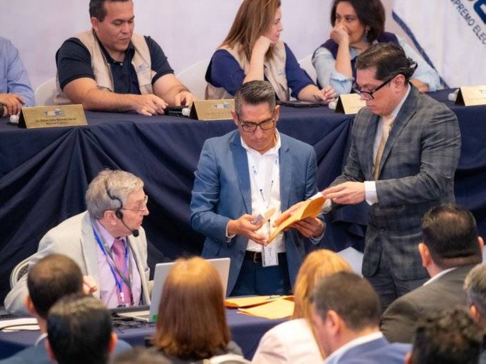 El secretario general del TSE, Erick Vides, a la derecha, entrega sobres manilas sellados con cinta adhesiva a los custodios de la urna digital. Le acompaña Luis Pardo, de Indra, a la izquierda. / TSE.,image_description: