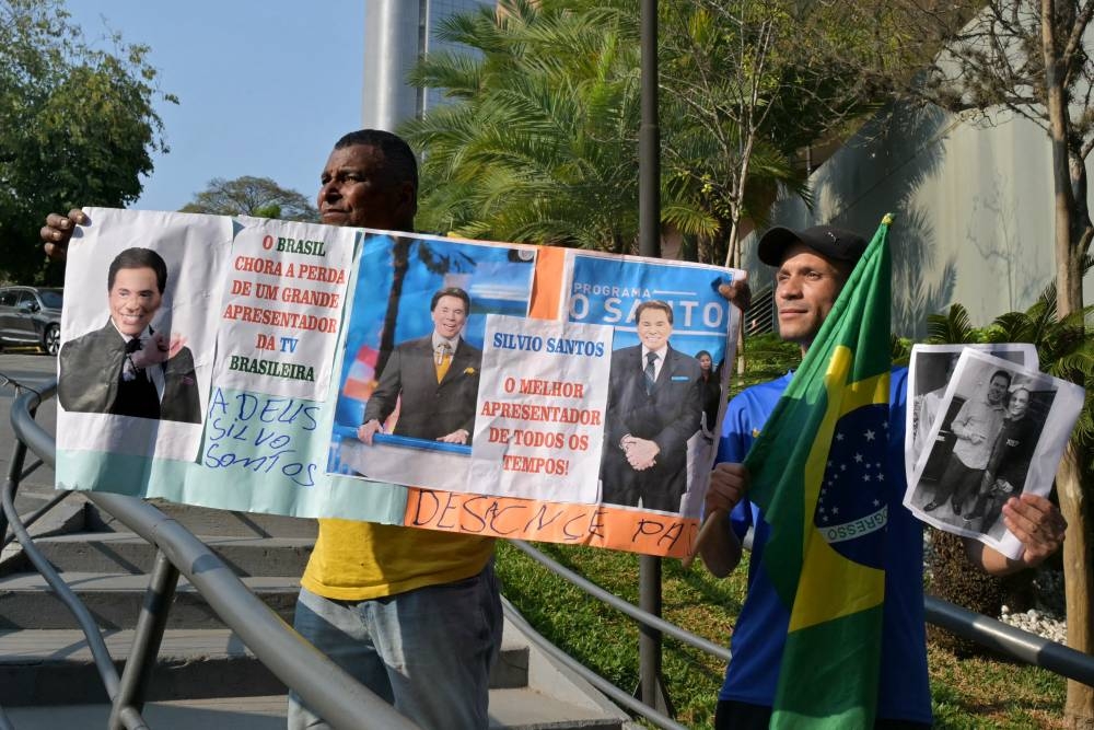 Dos hombres sostienen carteles de despedida en el exterior del Hospital Israelita Albert Einstein, donde falleció el presentador de televisión y empresario brasileño Senor Abravanel. /FP,image_description: