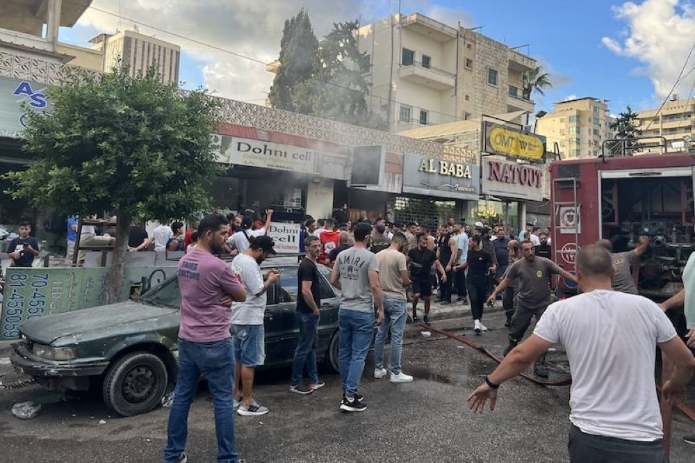 La gente se reúne mientras los bomberos apagan las llamas en el lugar de la explosión de un dispositivo en Saida, en el sur del Líbano este miércoles 18. Una segunda ola de explosiones de dispositivos mató a tres personas en bastiones de Hezbolá en el Líbano, lo que aumenta los temores de una guerra total entre Israel y los militantes respaldados por Irán./ AFP,image_description: