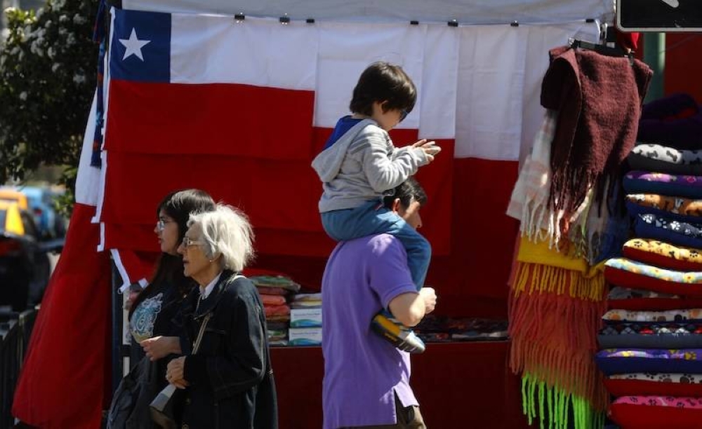 Un padre con su hijo en brazos pasa junto a banderas chilenas en venta en Santiago. En la última década, los nacimientos han caído un 29 por ciento en Chile, ahora el país con la tasa de natalidad más baja de América. / AFP,image_description: