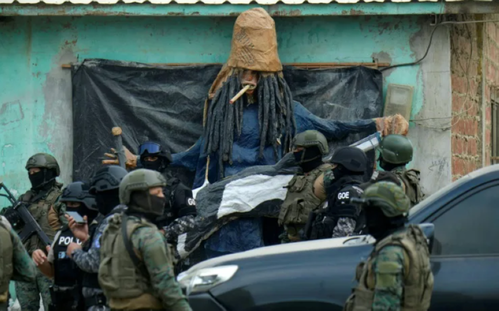 Personal de las fuerzas de seguridad pasa junto a un muñeco fabricado por miembros de la banda Latin King durante una operación conjunta de la Policía Nacional y las Fuerzas Armadas en un barrio de Durán, Ecuador / AFP.,image_description: