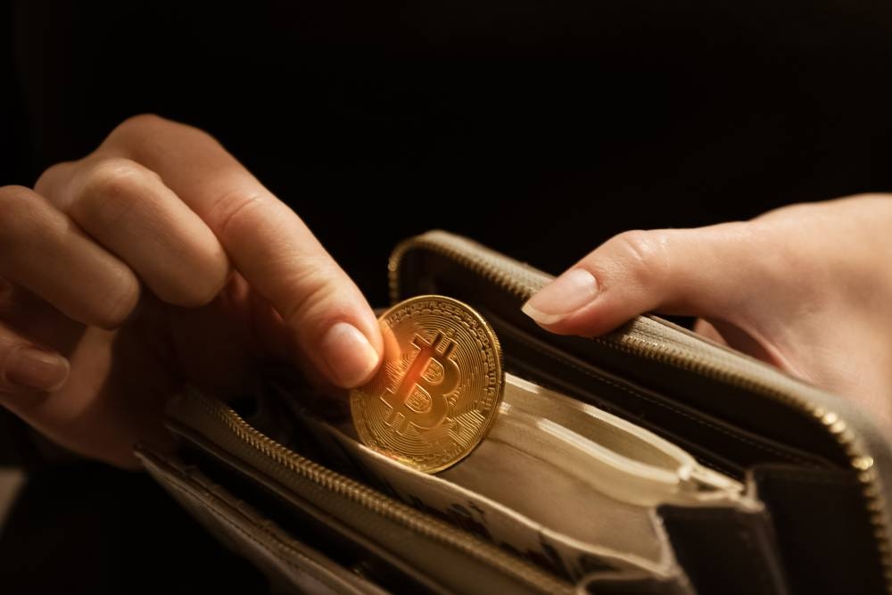 El bitcoin es la criptomoneda más popular del mercado y está muy cerca de tocar su máximo histórico. /DEM,image_description:Woman puts gold bitcoin coin in her purse, close up hands shot