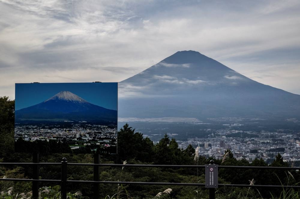 Un cartel con una foto del Monte Fuji cubierto de nieve se ve en un mirador mientras el Monte Fuji, la montaña más alta de Japón con 3.776 metros, se alza al fondo en Gotemba, prefectura de Shizuoka, este 31 de octubre de 2024. /AFP,image_description: