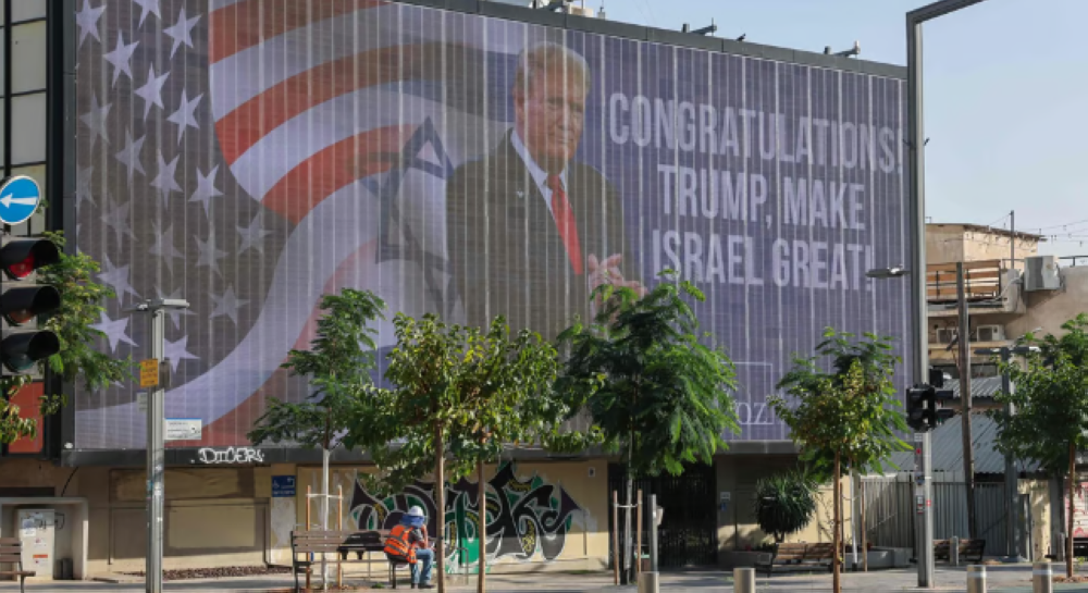 Un hombre sentado en un banco público debajo de una gran valla que felicita al presidente electo de Estados Unidos, Donald Trump, en Tel Aviv el miércoles/ Foto AFP.,image_description: