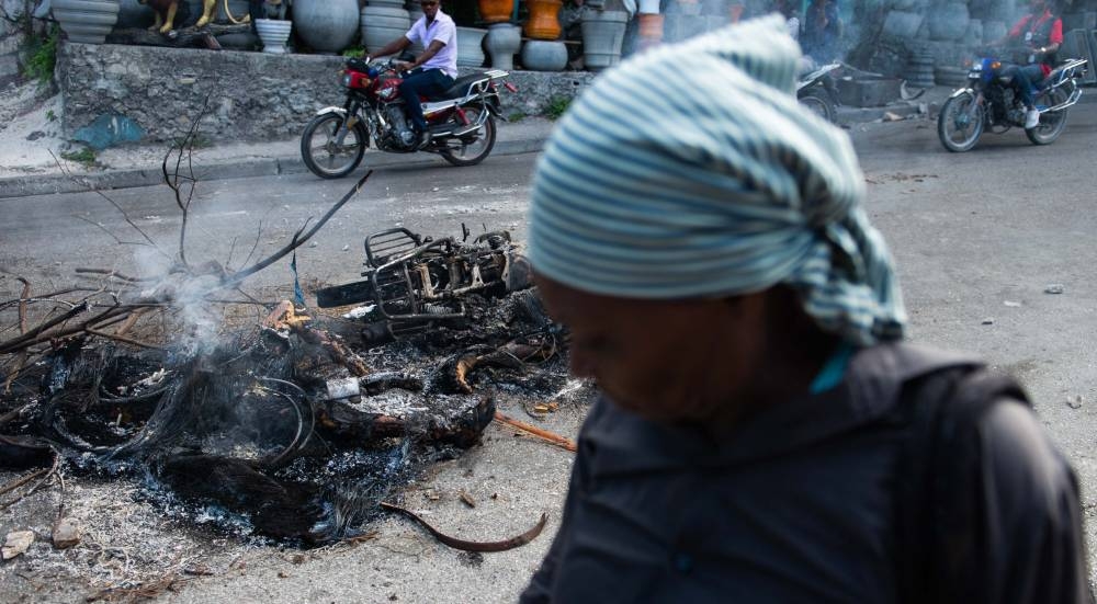 Personas pasan junto a los restos humeantes de los cuerpos de presuntos miembros de pandillas en PétionVille, un suburbio de Puerto Príncipe /Clarens Siffroy AFP.,image_description: