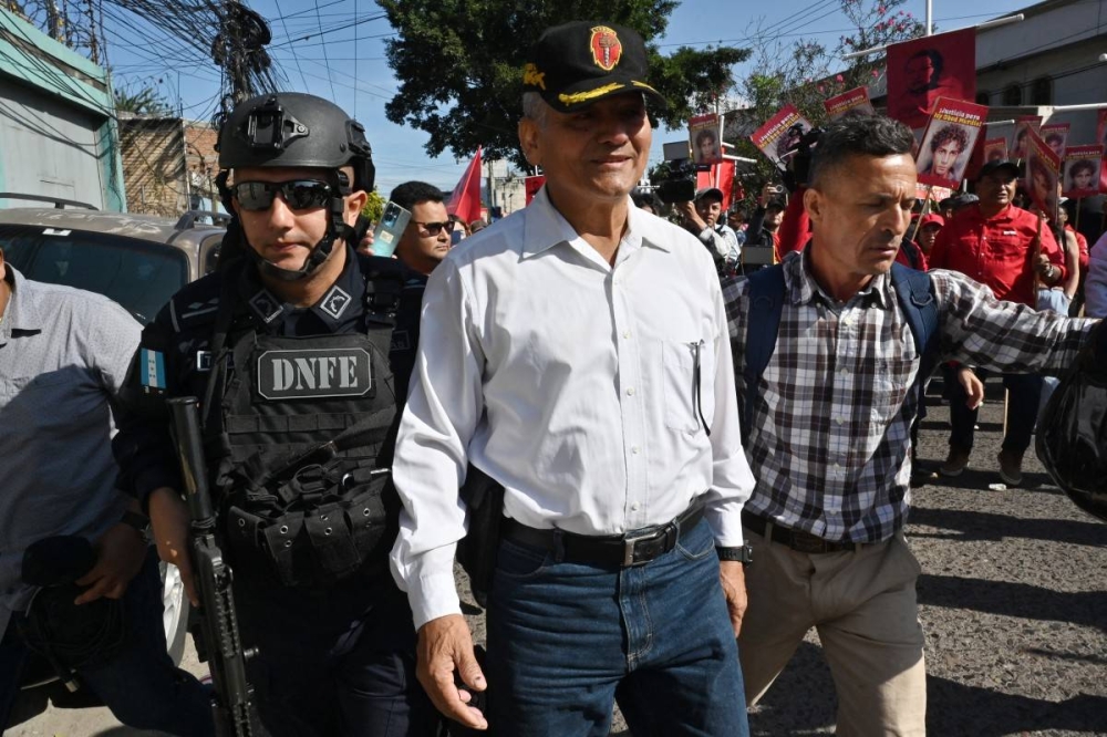 El coronel retirado Mario Maldonado Muñoz llega a la audiencia mientras simpatizantes del gobernante Partido Libertad y Refundación  realizan una manifestación exigiendo justicia para el exjefe del Estado Mayor Conjunto, general Romeo Vásquez Velásquez, durante la audiencia inicial por el asesinato de Isy Obed Murillo, en Tegucigalpa, el 10 de enero de 2025./ AFP,image_description: