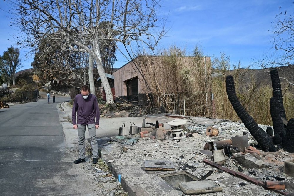 El arquitecto estadounidense Michael Kovac se encuentra cerca de los restos carbonizados de una casa vecina fuera de su casa (atrás), que sobrevivió al incendio Palisades debido a las características resistentes al fuego utilizadas en su construcción, en el vecindario Pacific Palisades de Los Ángeles./ Foto Frederic J. Brown AFP.